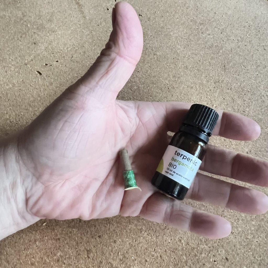 Fotografia dłoni trzymającej szklaną buteleczkę naturalnego, organicznego olejku eterycznego z bergamotki marki Terpenic obok małego stożka ziołowego do moksoterapii (moksy), zaprezentowana na prostym tle ze sklejki. Te elementy reprezentują naturalne metody walki ze stresem: aromaterapię kliniczną i moksoterapię.